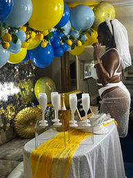 Bridal shower bachelorette scene: woman in white veil and lingerie beside an indoor table with champagne flutes and bottle in an ice bucket, yellow-and-blue lemon balloon garland, gold pillows, and festive accents.