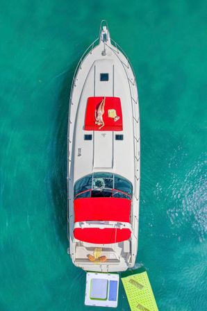 Aerial drone shot of a white luxury yacht anchored in turquoise water, featuring a red sun pad with a person sunbathing, beach towel and inflatable floating docks.