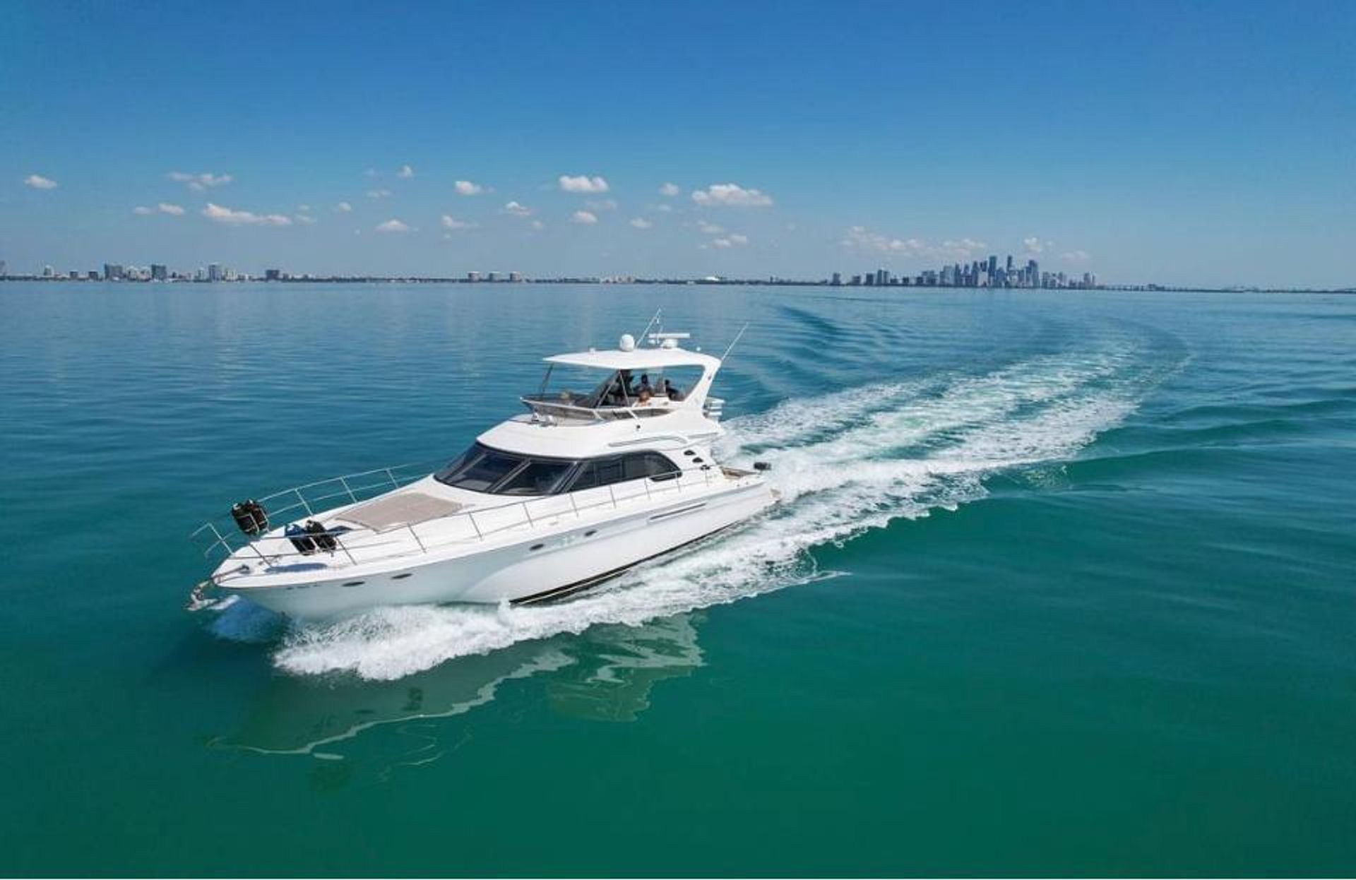 White luxury motor yacht cruising on calm turquoise water leaving a long wake, distant coastal city skyline on the horizon under a clear blue sky