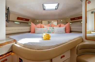 Cozy sunlit yacht v-berth cabin with cream leather trim, white quilted bed, coral and patterned throw pillows, overhead hatch skylight, wood accents and wall TV.
