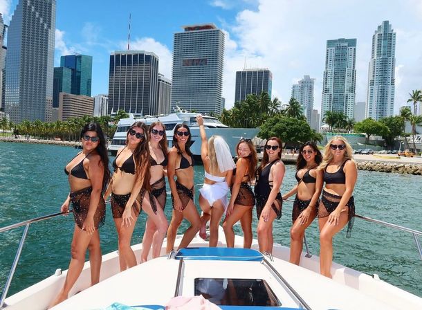 Bachelorette party on a Miami yacht — women in black swimsuits and a bride in a white veil posing on the bow with turquoise water, palm trees and downtown high-rise skyline under a sunny blue sky.