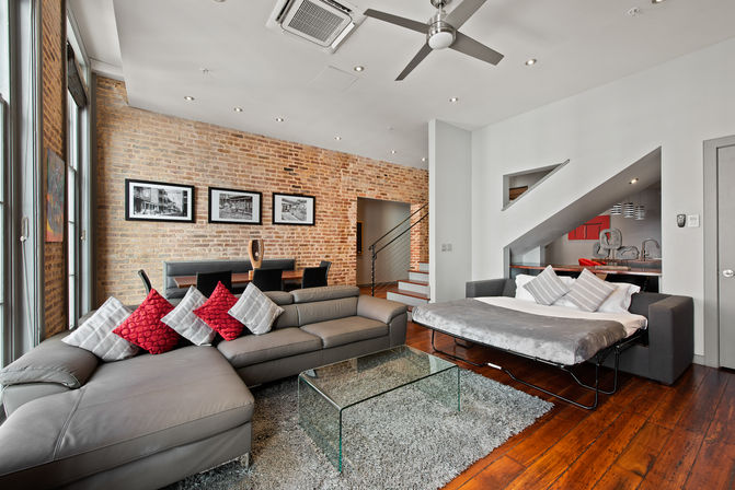 Cozy modern downtown loft living room with exposed brick wall, large windows, gray sectional with red accent pillows, glass coffee table, pull-out sofa bed, dining area and hardwood floors.