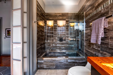 Contemporary spa-like bathroom with dark wood-look tile walls, glass-enclosed shower and built-in soaking tub, recessed shelf with black bottles, wall-mounted showerhead, lavender towel on a rail, and warm wooden vanity countertop.