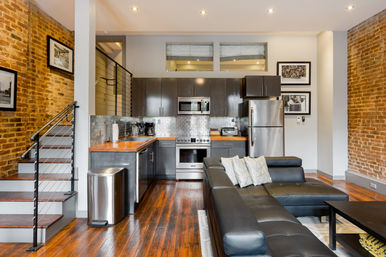 Industrial-chic open-concept loft kitchen and living room with exposed brick walls, hardwood floors, stainless steel appliances, dark cabinets, wooden countertops, subway-tile backsplash, metal cable staircase, and black leather sectional.