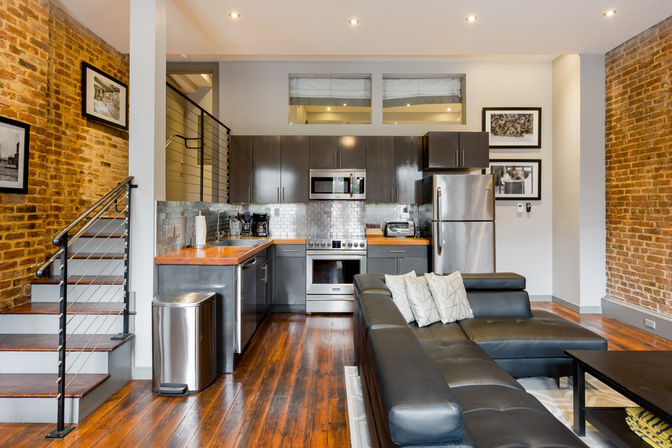 Industrial-chic open-concept loft kitchen and living room with exposed brick walls, hardwood floors, stainless steel appliances, dark cabinets, wooden countertops, subway-tile backsplash, metal cable staircase, and black leather sectional.