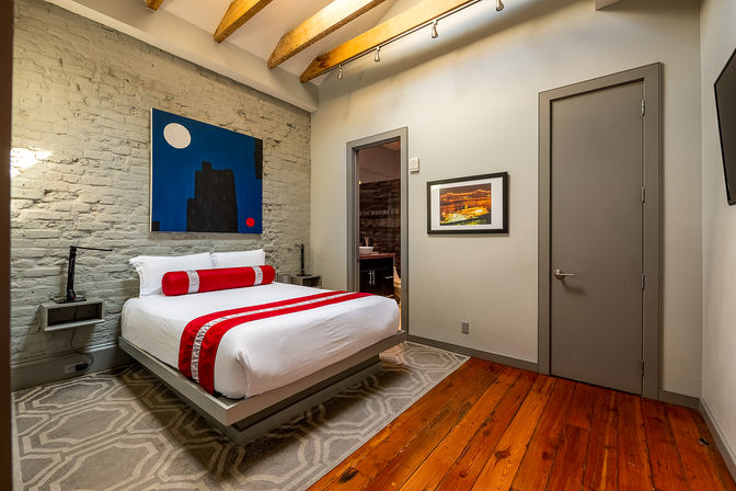 Modern urban loft bedroom with floating platform bed in white linens and red accent runner and bolster, painted brick feature wall, wooden ceiling beams, hardwood floors, abstract wall art and doorway to en-suite bathroom.