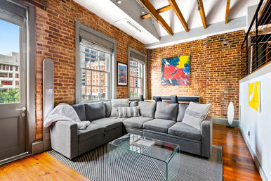 Sunny urban loft living room with exposed brick walls and wooden beams, large windows with city views, a gray sectional sofa, glass coffee table on a patterned rug, hardwood floors and colorful abstract wall art.