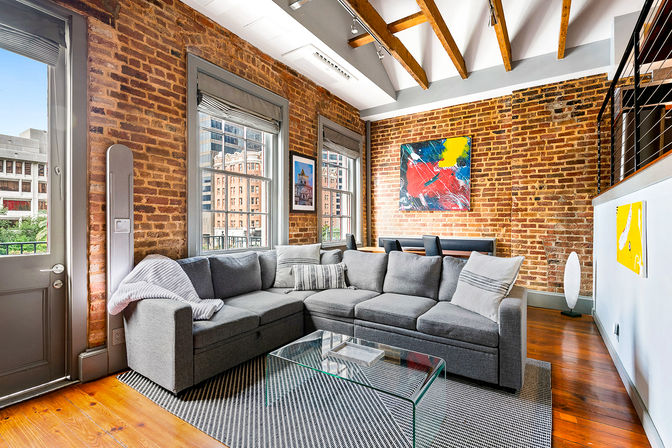Sunny urban loft living room with exposed brick walls and wooden beams, large windows with city views, a gray sectional sofa, glass coffee table on a patterned rug, hardwood floors and colorful abstract wall art.