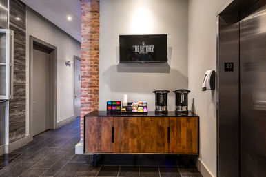 Cozy self-service coffee station in a modern hotel hallway — two urns on a wooden cabinet, colorful coffee pods, exposed brick column and nearby elevator.