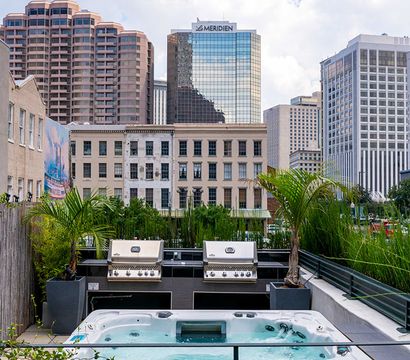 Rooftop hot tub and twin stainless-steel grills on an urban terrace with potted palms and a downtown high-rise skyline under a bright blue sky.