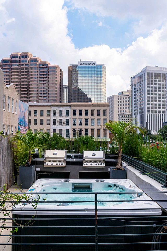 Rooftop hot tub and twin stainless-steel grills on an urban terrace with potted palms and a downtown high-rise skyline under a bright blue sky.