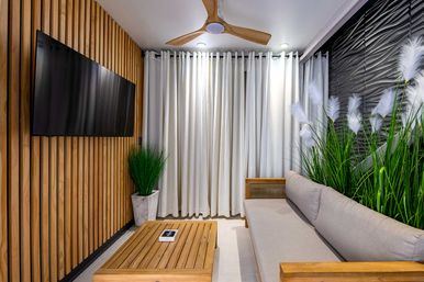 Modern living room interior with wooden slat accent wall and wall-mounted flat-screen TV, neutral gray sofa and wooden coffee table, tall indoor grasses, white floor-to-ceiling curtains and a wooden ceiling fan