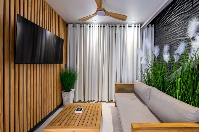 Modern living room interior with wooden slat accent wall and wall-mounted flat-screen TV, neutral gray sofa and wooden coffee table, tall indoor grasses, white floor-to-ceiling curtains and a wooden ceiling fan