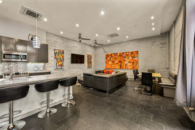 Modern urban loft: open kitchen with stainless appliances and black bar stools, gray leather sectional, vibrant orange abstract art on white painted brick, and dining banquette.
