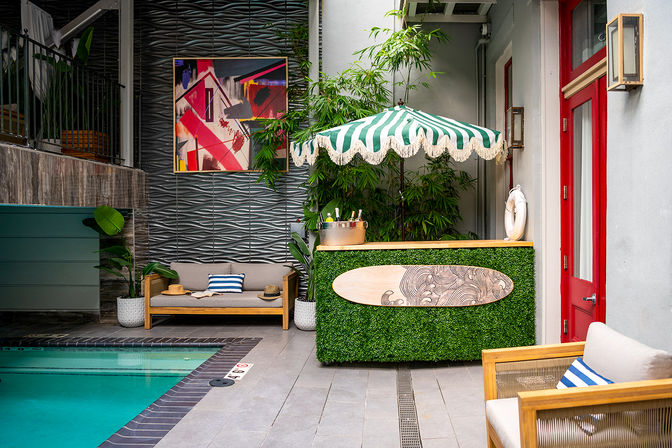 Boutique hotel poolside courtyard with turquoise swimming pool, green-and-white striped umbrella over a boxwood-clad bar with surfboard sign, tropical plants, abstract wall art and cozy wooden lounge seating.