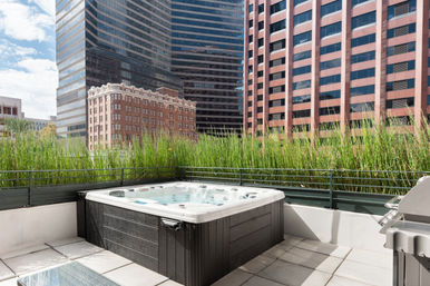 Sunny rooftop hot tub on a downtown terrace — an urban oasis framed by tall grasses and surrounded by reflective glass and brick skyscrapers.