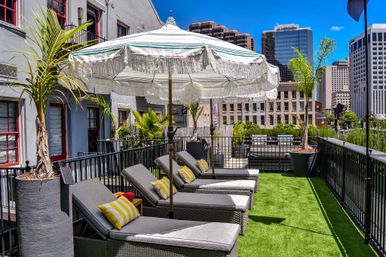 Sunny urban rooftop terrace with cushioned lounge chairs, striped fringed umbrella, potted palms and artificial turf overlooking a downtown high-rise skyline.