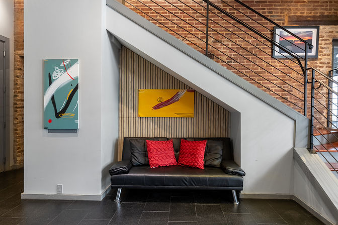 Cozy under-stair nook with a black leather sofa and two red throw pillows beneath a modern metal staircase in an industrial loft entryway, exposed brick, wood-slat accent wall and colorful contemporary art.