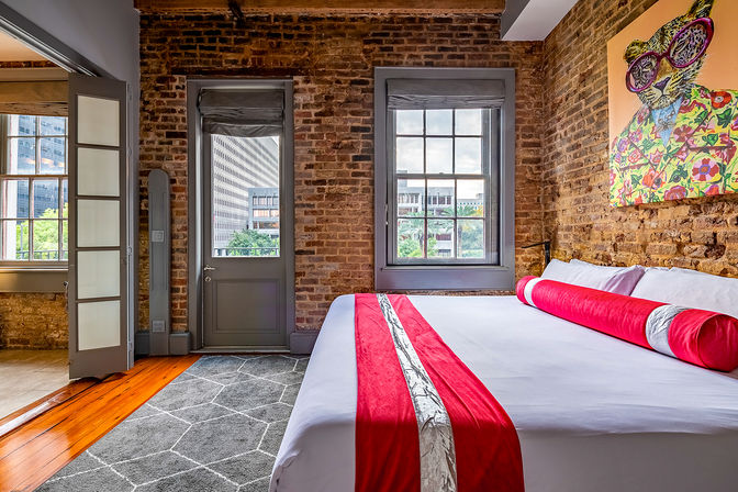 Urban loft bedroom with exposed brick walls, king bed with white linens and red runner, playful pop-art leopard painting and city-view windows.