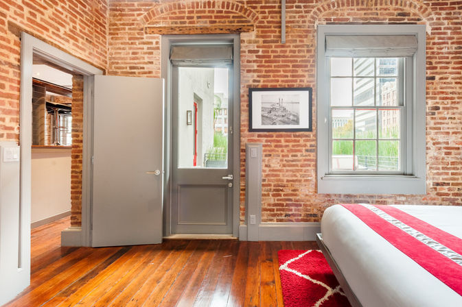 Cozy urban loft bedroom with exposed brick walls and polished hardwood floors, gray door and window revealing city buildings, bed with red-striped bedding and matching red rug