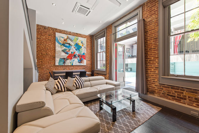 Bright urban loft living room with exposed brick walls, beige leather L-shaped sectional with striped pillows, glass coffee table, vibrant abstract painting, large windows and door opening to a sunny courtyard/pool and a small dining area — industrial-chic interior.
