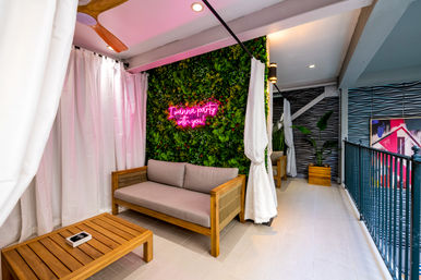 Cozy modern indoor balcony lounge with wooden sofa and coffee table, lush vertical green wall, pink neon sign reading 'I wanna party with you', white curtains and potted plants.
