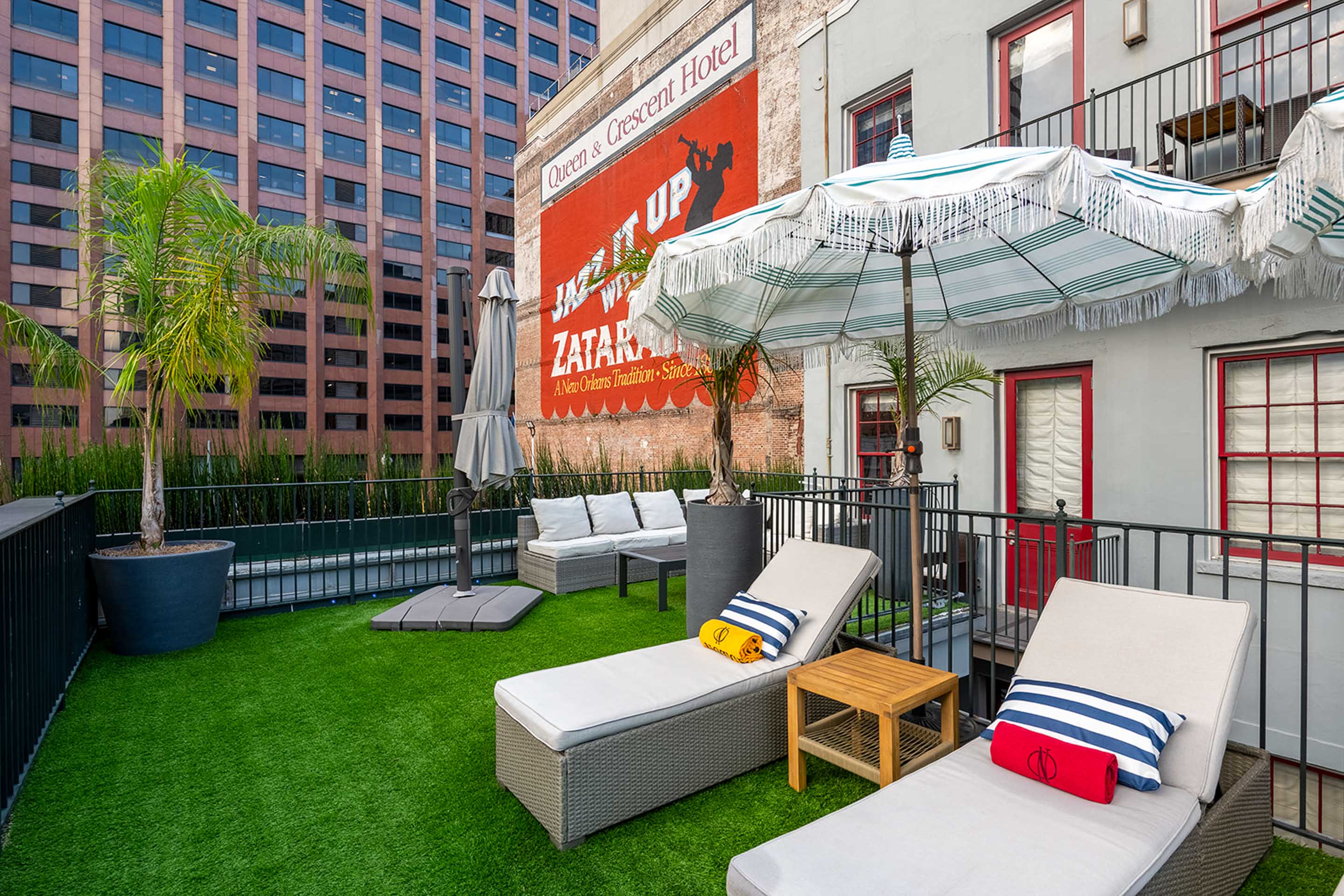 Sunny urban rooftop patio with cushioned lounge chairs, striped fringe umbrellas, potted palm trees and bright artificial turf beside a brick wall featuring a vintage painted advertisement and tall office buildings