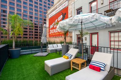 Sunny urban rooftop patio with cushioned lounge chairs, striped fringe umbrellas, potted palm trees and bright artificial turf beside a brick wall featuring a vintage painted advertisement and tall office buildings