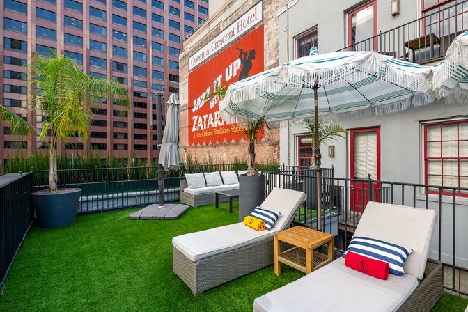 Sunny urban rooftop patio with cushioned lounge chairs, striped fringe umbrellas, potted palm trees and bright artificial turf beside a brick wall featuring a vintage painted advertisement and tall office buildings