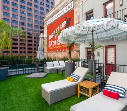 Sunny urban rooftop patio with cushioned lounge chairs, striped fringe umbrellas, potted palm trees and bright artificial turf beside a brick wall featuring a vintage painted advertisement and tall office buildings