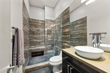 Sleek spa-like modern bathroom interior with glass-enclosed walk-in shower, wood-look tile and built-in bench, white toilet, round vessel sink on dark vanity, gray towels.