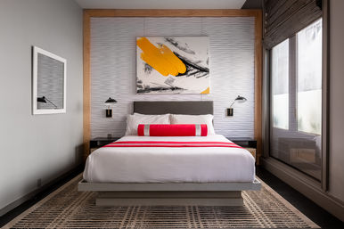 Stylish urban boutique hotel room with king bed, crisp white linens, red runner and bolster, abstract yellow-and-black painting above gray headboard, wall-mounted reading lamps and large window with gray shades