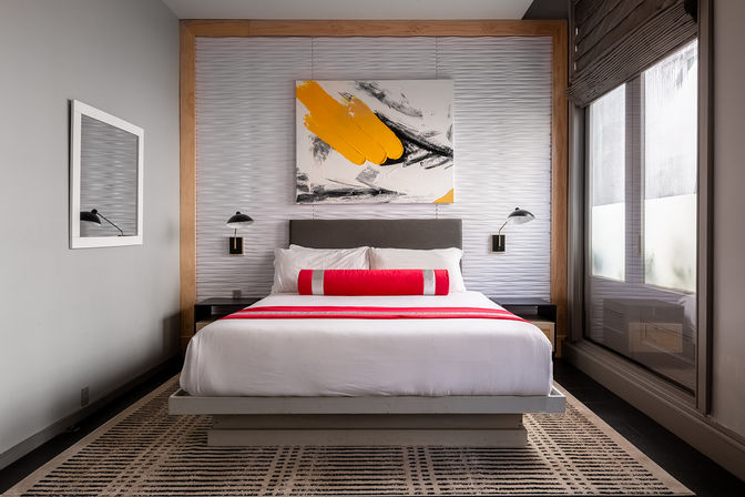 Stylish urban boutique hotel room with king bed, crisp white linens, red runner and bolster, abstract yellow-and-black painting above gray headboard, wall-mounted reading lamps and large window with gray shades