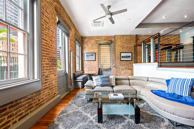 Industrial-chic urban loft living room with exposed brick walls, tall windows with city views, curved light-gray leather sectional with blue striped pillows, glass coffee table on a gray shag rug, hardwood floors and metal mezzanine railing.