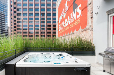 Cozy rooftop hot tub on an urban terrace in New Orleans with tall ornamental grasses, stainless-steel grill, red brick mural advertising a local tradition, and surrounding high-rise office buildings.