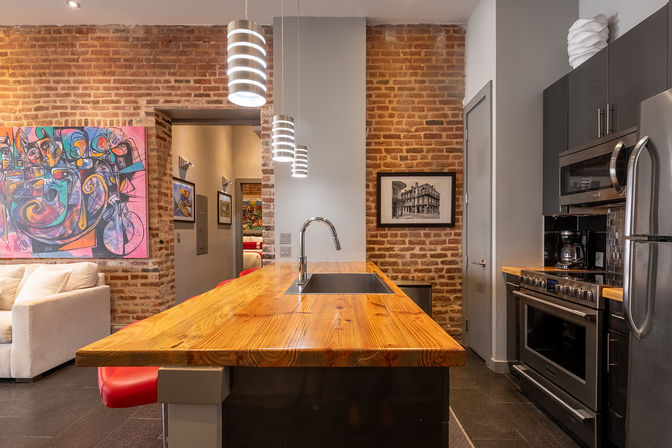 Cozy urban loft kitchen with exposed brick walls, a wide wooden island with sink, stainless steel appliances, pendant lights, and colorful abstract wall art.