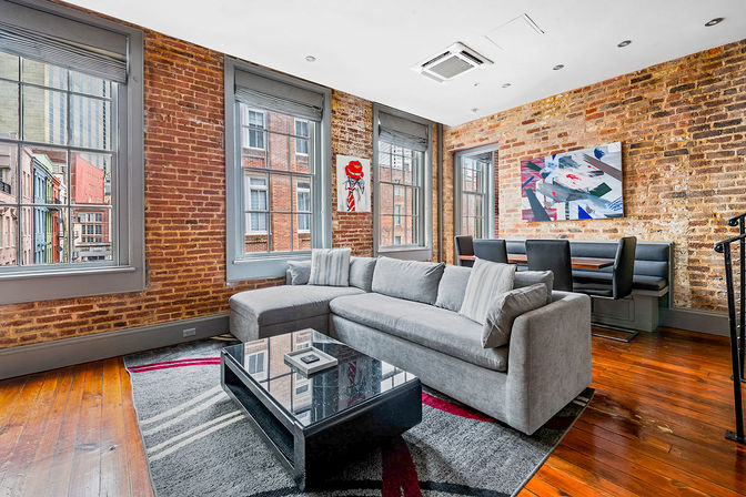 Bright industrial loft living room with exposed brick walls, hardwood floors, large city-view windows, a modern gray sectional, black glass coffee table, built-in banquette dining, and colorful wall art.