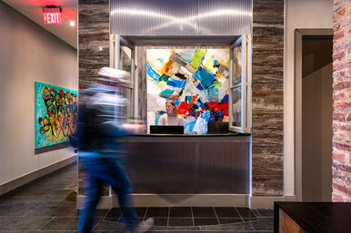 Modern hotel lobby reception with vibrant abstract paintings behind the front desk, a receptionist at the window and a blurred guest walking by, exposed brick and dark tile floor.