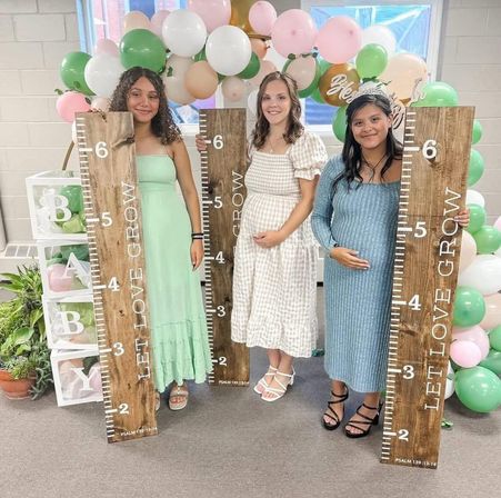 Three pregnant women at an indoor baby shower holding rustic wooden growth rulers reading “LET LOVE GROW,” with pastel pink, green and white balloons, white BABY blocks, and smiling, cradling their baby bumps — maternity celebration with rustic party decor.