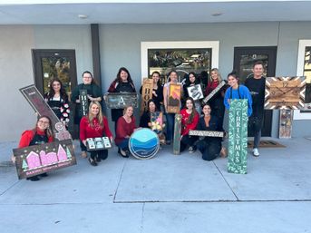Smiling group of about 15 adults posed outside a craft studio, each holding handmade wooden signs and décor — holiday and home-themed pieces like a Christmas plank, baking sign, painted clock and colorful DIY wood projects from a workshop.