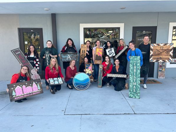 Smiling group of about 15 adults posed outside a craft studio, each holding handmade wooden signs and décor — holiday and home-themed pieces like a Christmas plank, baking sign, painted clock and colorful DIY wood projects from a workshop.
