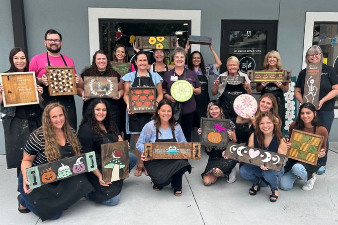 Smiling group of adults posing outside a storefront after a paint-and-craft workshop, each holding handmade wooden signs featuring pumpkins, moons, florals and other seasonal designs.