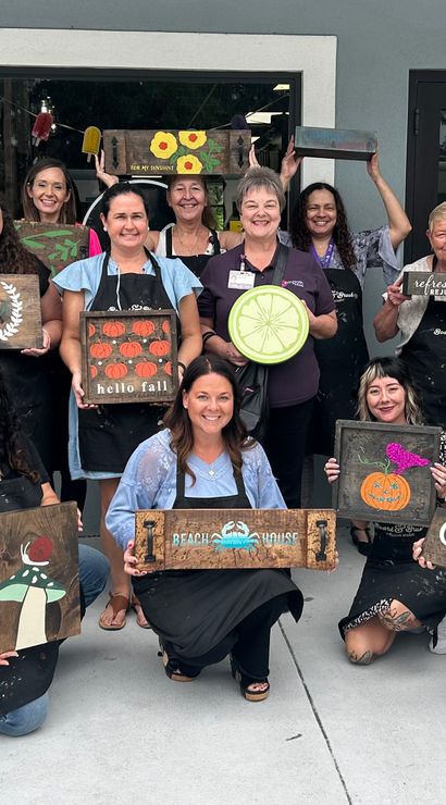 Smiling group of adults posing outside a storefront after a paint-and-craft workshop, each holding handmade wooden signs featuring pumpkins, moons, florals and other seasonal designs.
