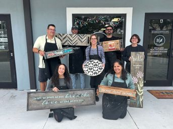 Seven adults smiling outside a craft studio entrance holding painted rustic wooden signs and a round geometric board — group photo from a DIY wood sign workshop.