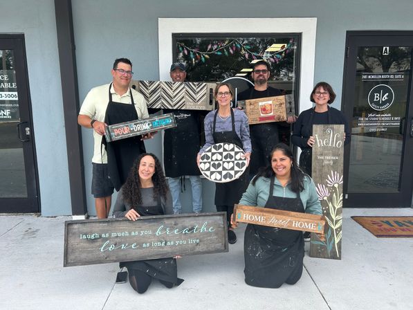 Seven adults smiling outside a craft studio entrance holding painted rustic wooden signs and a round geometric board — group photo from a DIY wood sign workshop.