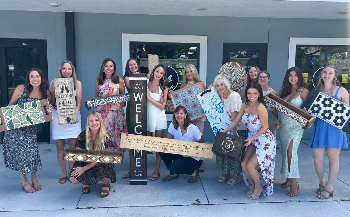 Smiling group of women posing outside a storefront on a sunny day, each holding colorful handmade wooden signs and decorative trays with welcome, floral and geometric home‑decor designs — lively DIY craft gathering.