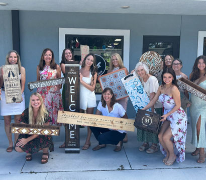 Smiling group of women posing outside a storefront on a sunny day, each holding colorful handmade wooden signs and decorative trays with welcome, floral and geometric home‑decor designs — lively DIY craft gathering.
