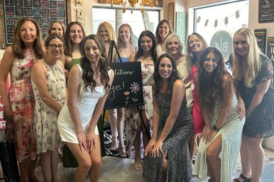 Smiling group of women in sundresses gathered indoors around a chalkboard easel with a floral “congratulations” sign, posing for a celebratory group photo in a bright boutique event space.