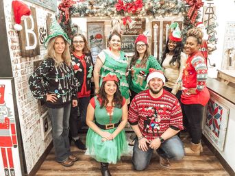 Smiling group of nine adults in ugly Christmas sweaters, elf costumes and Santa hats posing inside a cozy, decorated holiday shop with garlands, wreaths and festive signs — cheerful Christmas team photo.