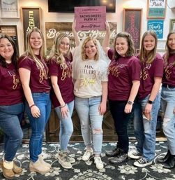 Group of seven friends at an indoor DIY wood‑sign workshop for a bachelorette party — bride in a veil and white tee holding a pink sign reading BACHELORETTE PARTY, bridesmaids in matching maroon shirts and jeans in front of a rustic wood sign wall.